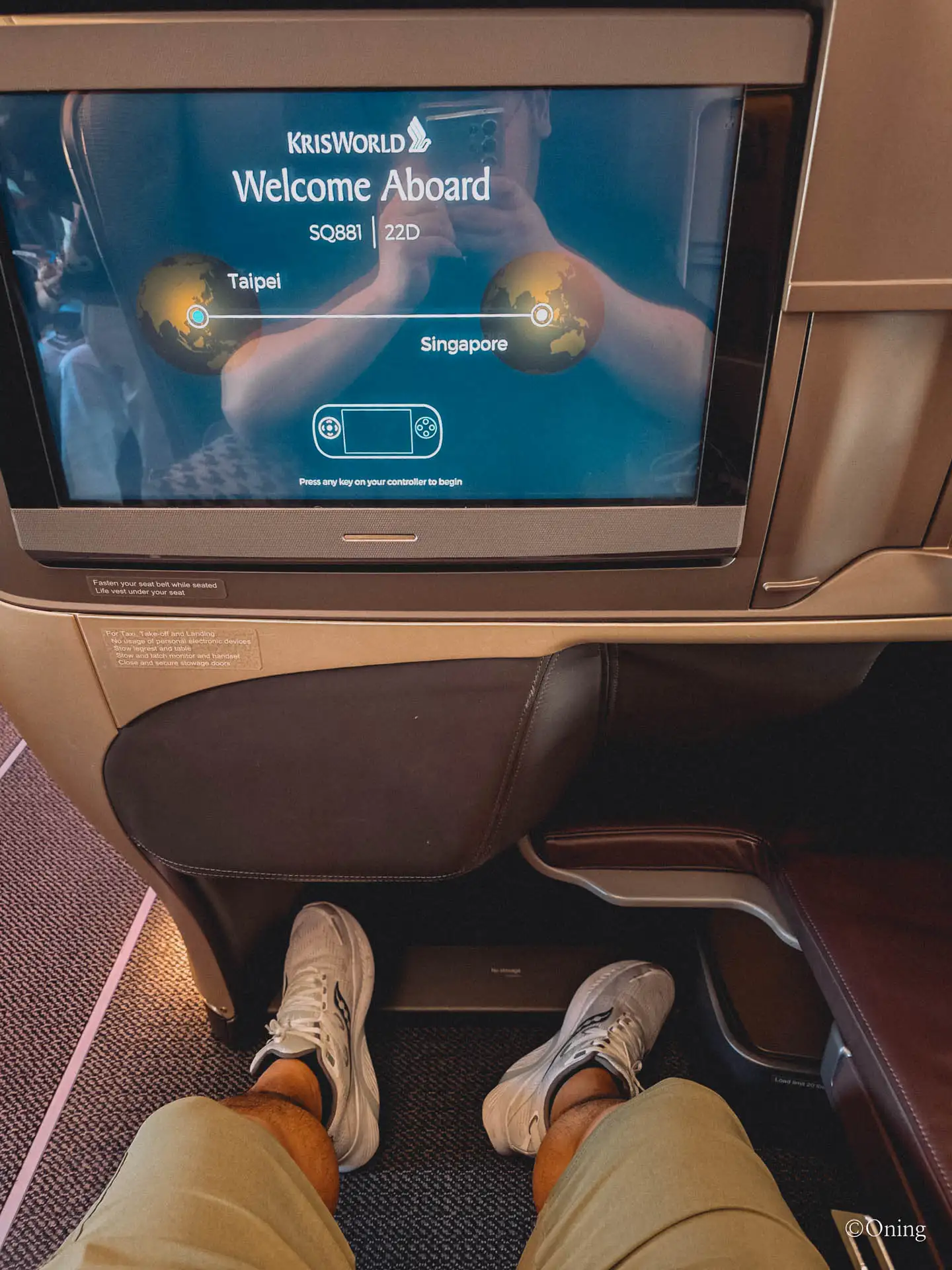 The footwell of a Singapore Airlines business class seat set diagonally to one side, with the KrisWorld welcome screen for SQ881 above — an angled lie-flat recliner rather than a full-flat bed