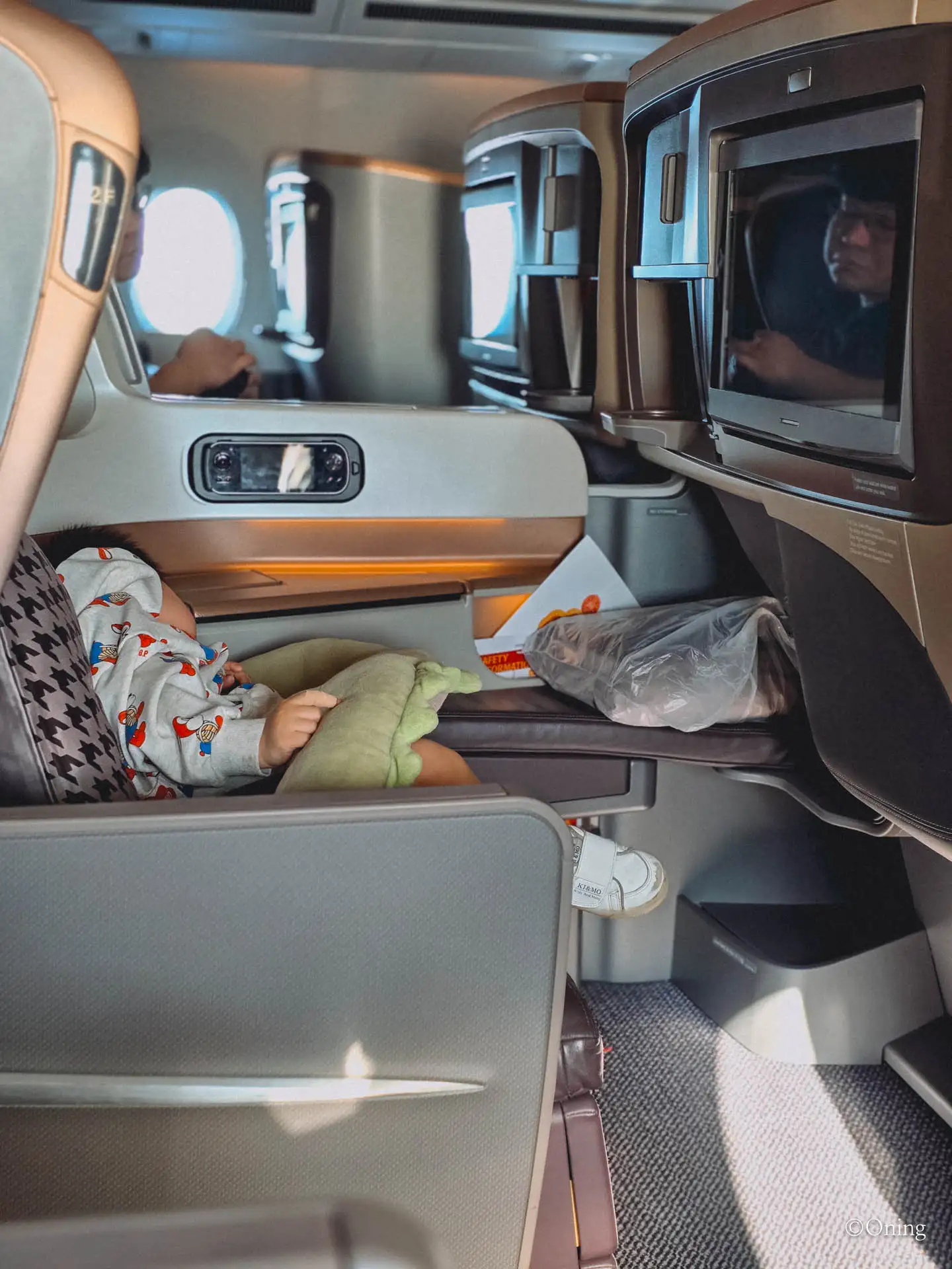 A toddler asleep and reclined under a blanket with a green dinosaur pillow in business class